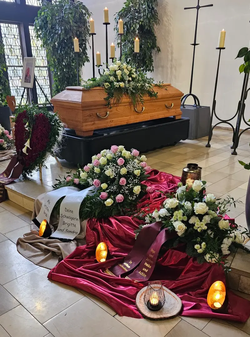 Ruhiger Abschiedsraum mit braunem Sarg, weißen Blumen, roten Tüchern und Kerzen